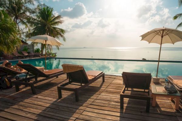 An inviting poolside with lounge chairs, umbrellas, a wooden deck, and a stunning view of the sea under a partly cloudy sky and palm trees.