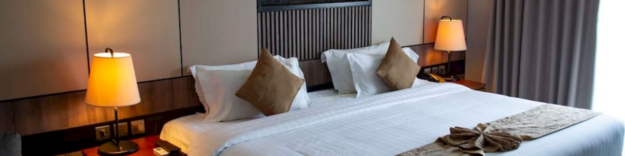 A neatly arranged hotel room with a large bed, two bedside tables with lamps, a wall-mounted art piece, and a bright window with curtains.