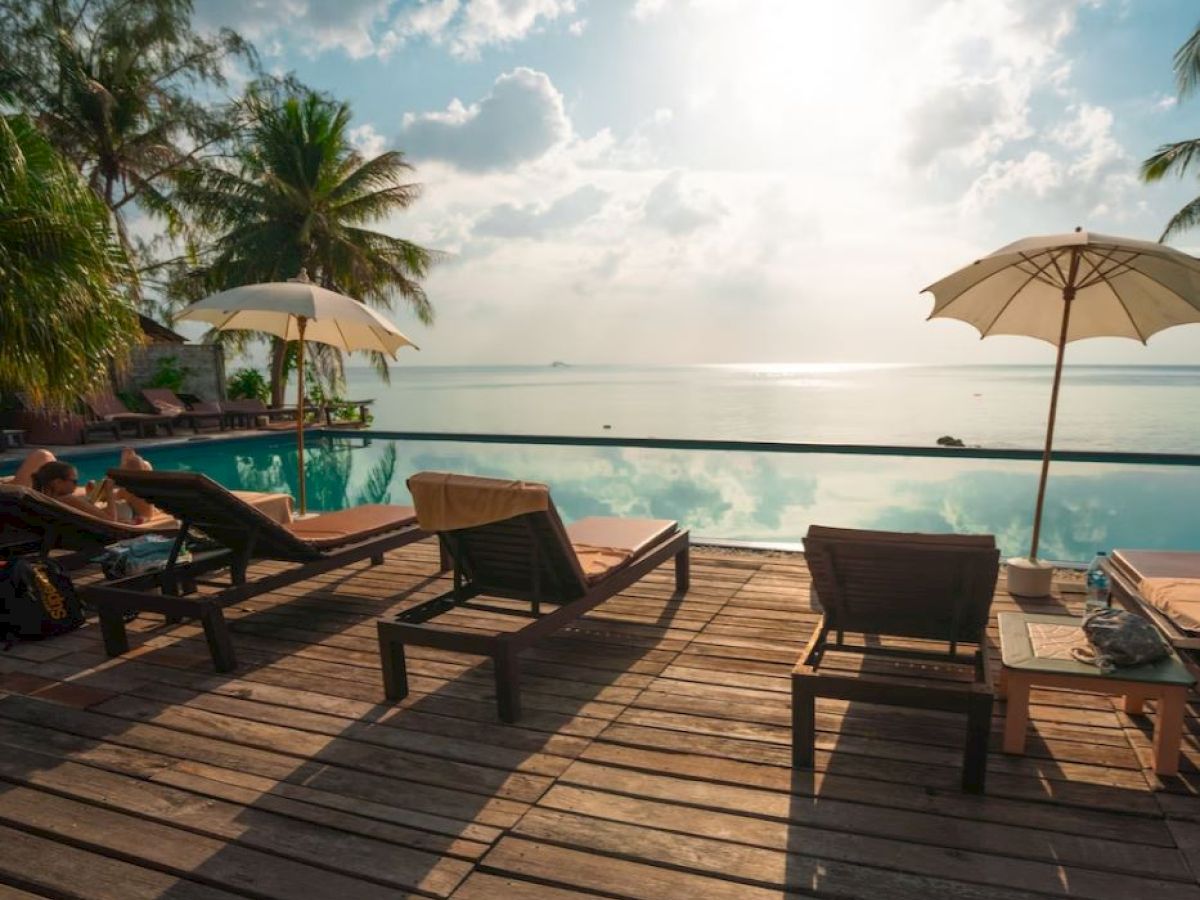 Poolside area with lounge chairs, umbrellas, and a stunning ocean view. Palms in the background, serene and sunny environment always ending the sentence.