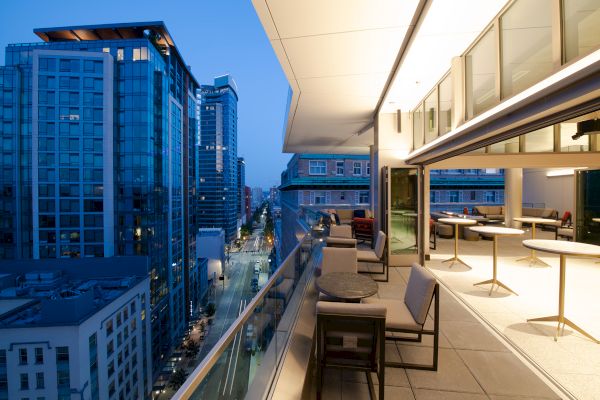 The image shows a modern cityscape at dusk, viewed from a balcony with a seating area and tables, overlooking buildings and a street below.