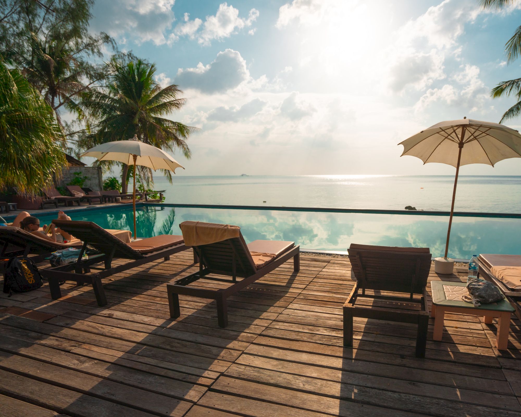 A serene poolside scene with lounge chairs, umbrellas, palm trees, and a reflective body of water beneath a partly cloudy sky during sunset.