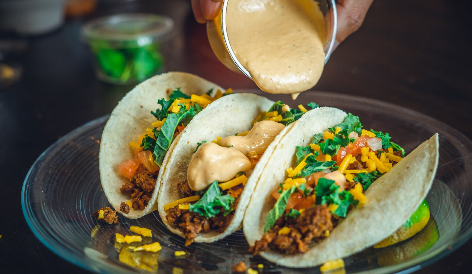 Three tacos filled with ground meat, cheese, kale, and tomatoes on a plate, with sauce being poured on top of them from a small container.