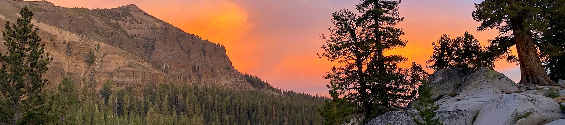 A serene landscape with a lake, trees, rocky terrain, and a mountain under a colorful sunset sky is depicted in this image.