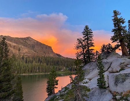 A serene landscape with a lake, trees, rocky terrain, and a mountain under a colorful sunset sky is depicted in this image.