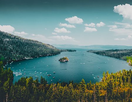 A scenic lake surrounded by dense forested hills, with an island in the center, under a partly cloudy sky, creating a beautiful landscape scene.