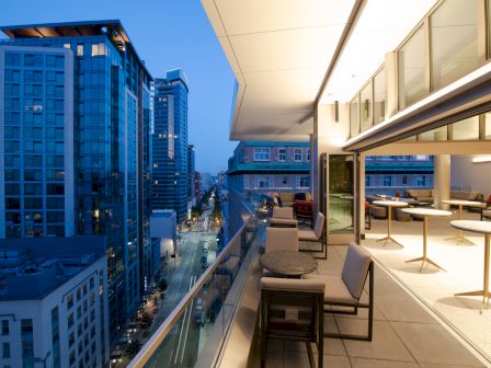 A modern outdoor terrace with chairs and tables overlooks a city street, flanked by tall buildings at dusk.