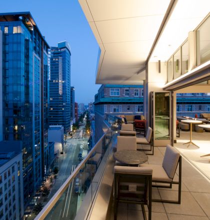 A modern cityscape at dusk, with a balcony featuring chairs and tables overlooking a street lined with high-rise buildings and lit windows.